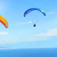 Two bright paragliders floating in a blue sky over the ocean.